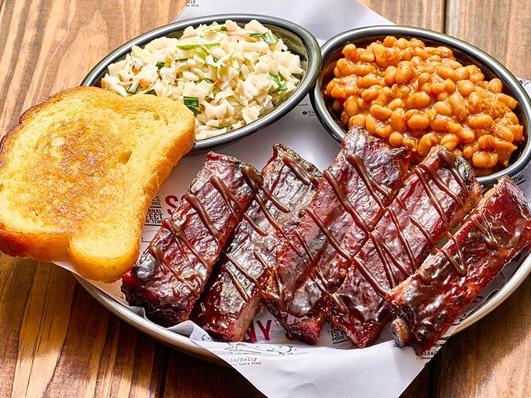 Sonny's BBQ slow-smoked St. Louis ribs with Sweet and Smokey glaze served on a platter with BBQ baked beans, coleslaw, and toasted garlic bread
